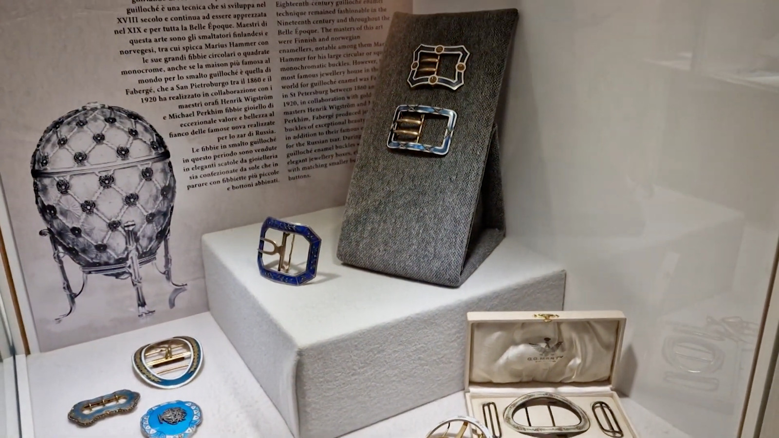 Guilloché enamel buckles on a grey felt wedge in front of a bilingual exhibition panel, display design by Sogand Nobahar