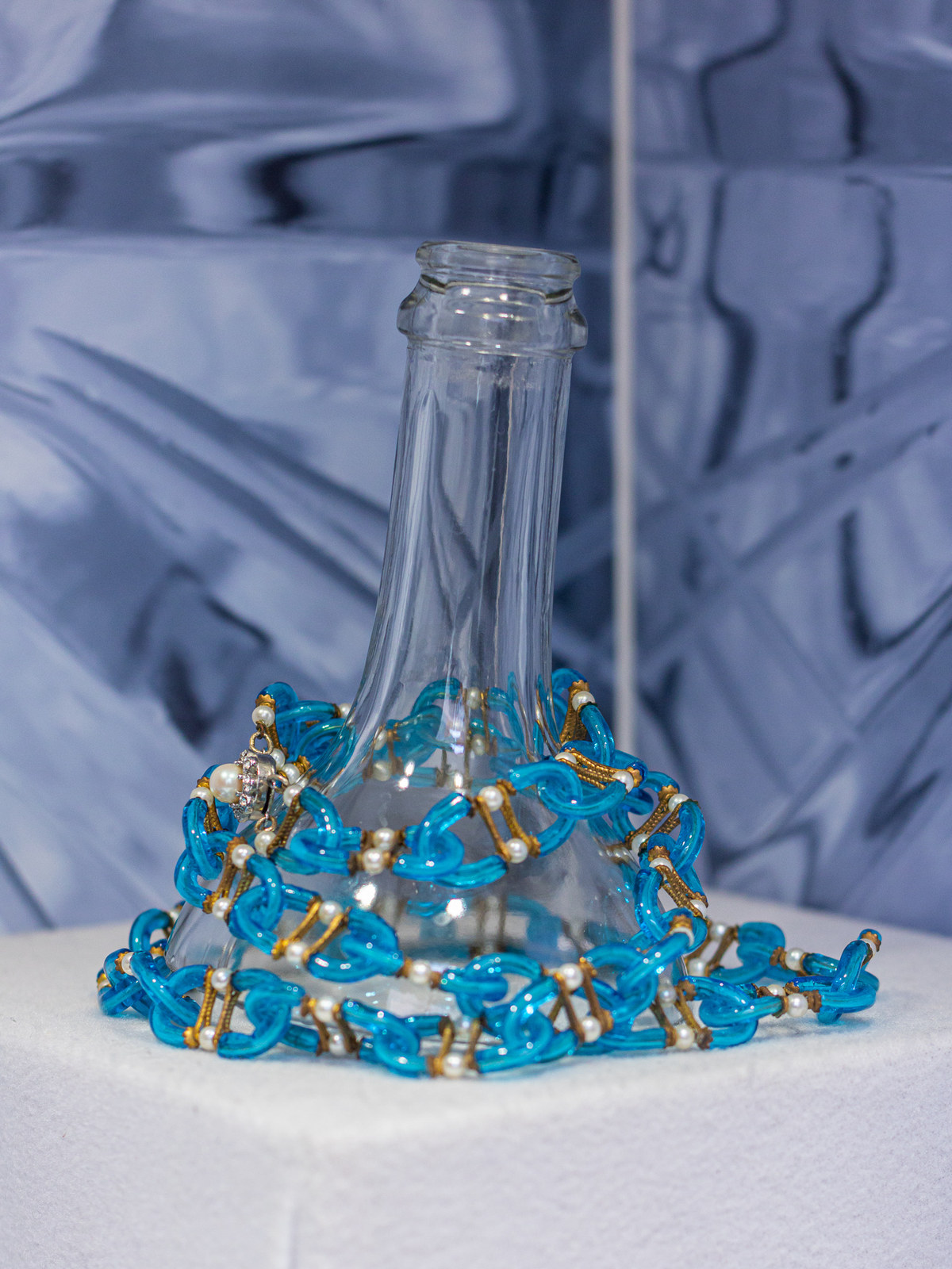 Blue glass chain necklace displayed around a cut bottle base against a macro glass background, Museo del Bijou