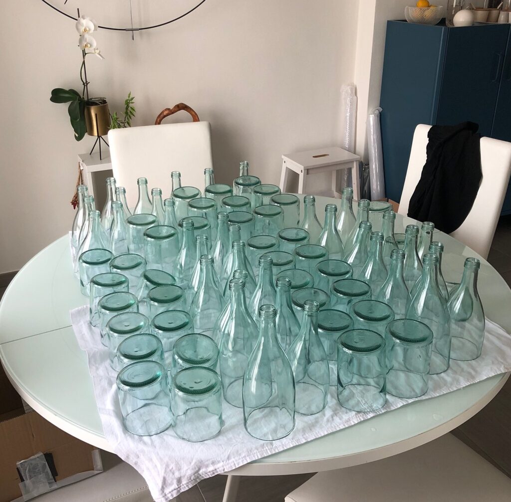 Sixty recovered glass bottles collected on a table before cutting, exhibition display material gathered by Sogand Nobahar