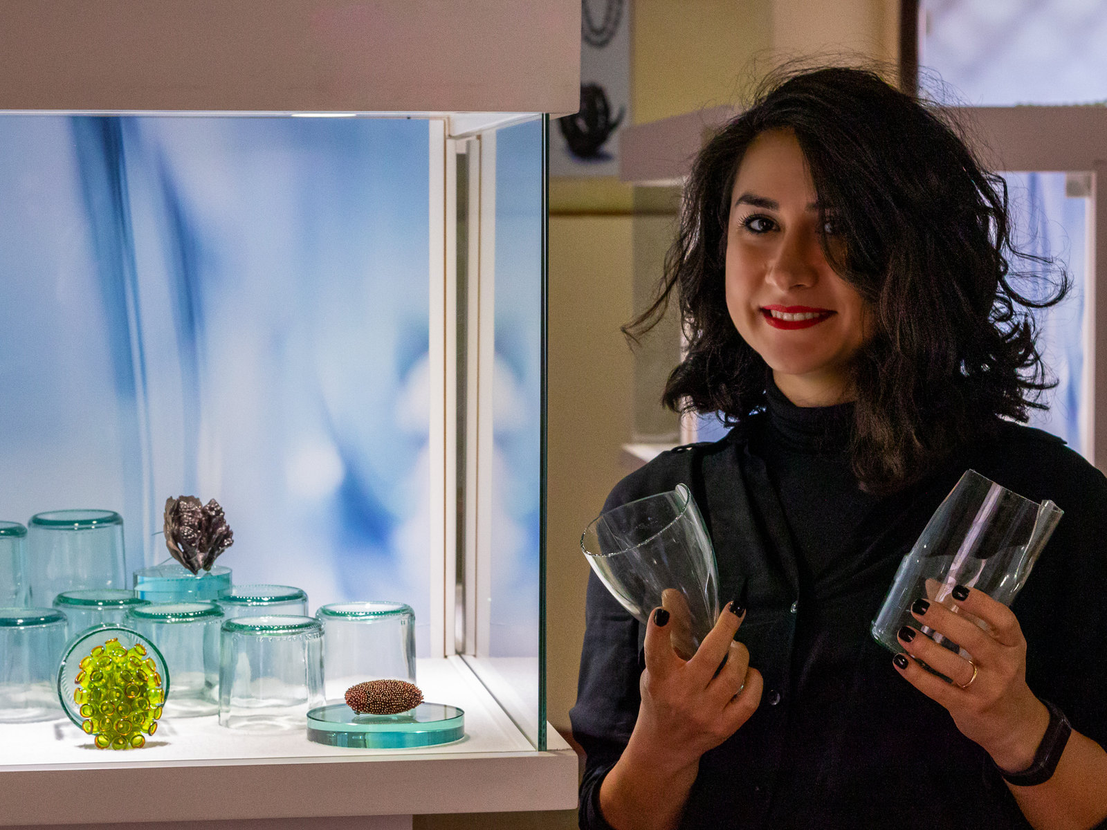 Sogand Nobahar holding two cut glass vessels beside the exhibition vitrine she designed