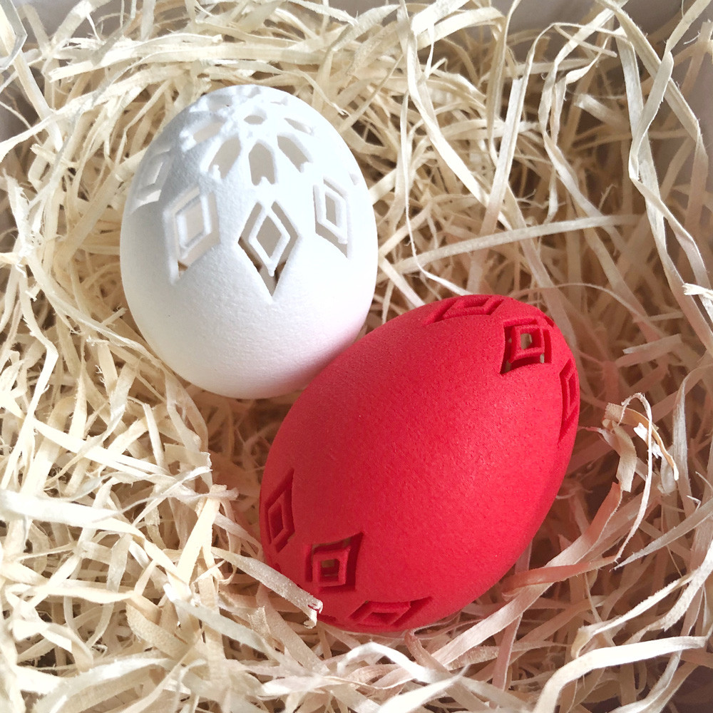 Two 3D printed Fertility Eggs, one white with cut-out geometric pattern and one red with raised relief, resting in wood wool, decorative objects by Nobahar Design
